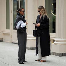 Anouk Yve looking at her phone as she and her friend stand together on a New York street wearing chic, neutral fall outfits.