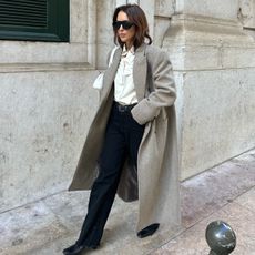 Debora wears a gray coat, white blouse, black pants, black sunglasses, a white bag, and black flats.