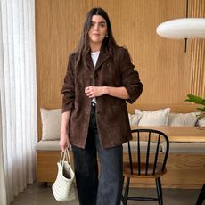 British fashion influencer Anna Newton poses in her modern London kitchen with wood panelling wearing a brown suede jacket, white t-shirt, gray baggy jeans, and a white woven tote bag