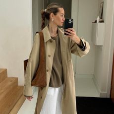 Dutch fashion influencer Anouk Yve wearing an oversize trench coat, a tan sweater, white linen pants, and suede shoulder bag