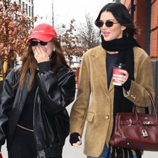 Hailey Bieber wears a red hat, black bomber jacket, black top, and black pants. Kendall Jenner wears a black scarf, black top, brown suede blazer, burgundy hermes bag, jeans, and loafers.